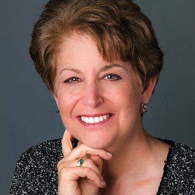 A portrait of Dr. Diane smiling with her right hand on her right cheek. She has short brown hair and is wearing a black and white speckled blouse.